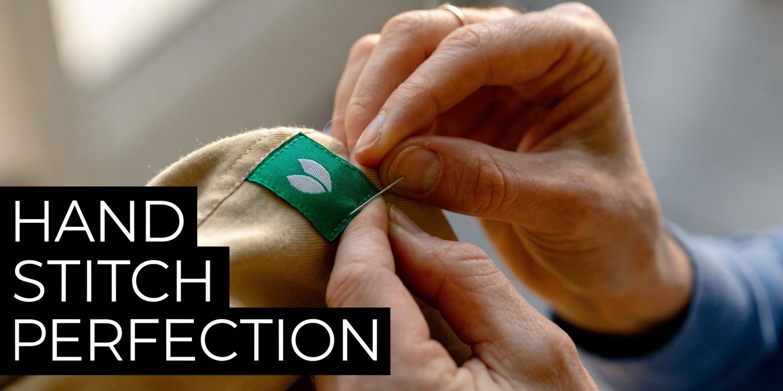 A close-up of a person's hands carefully hand stitching a small green label onto khaki clothing.